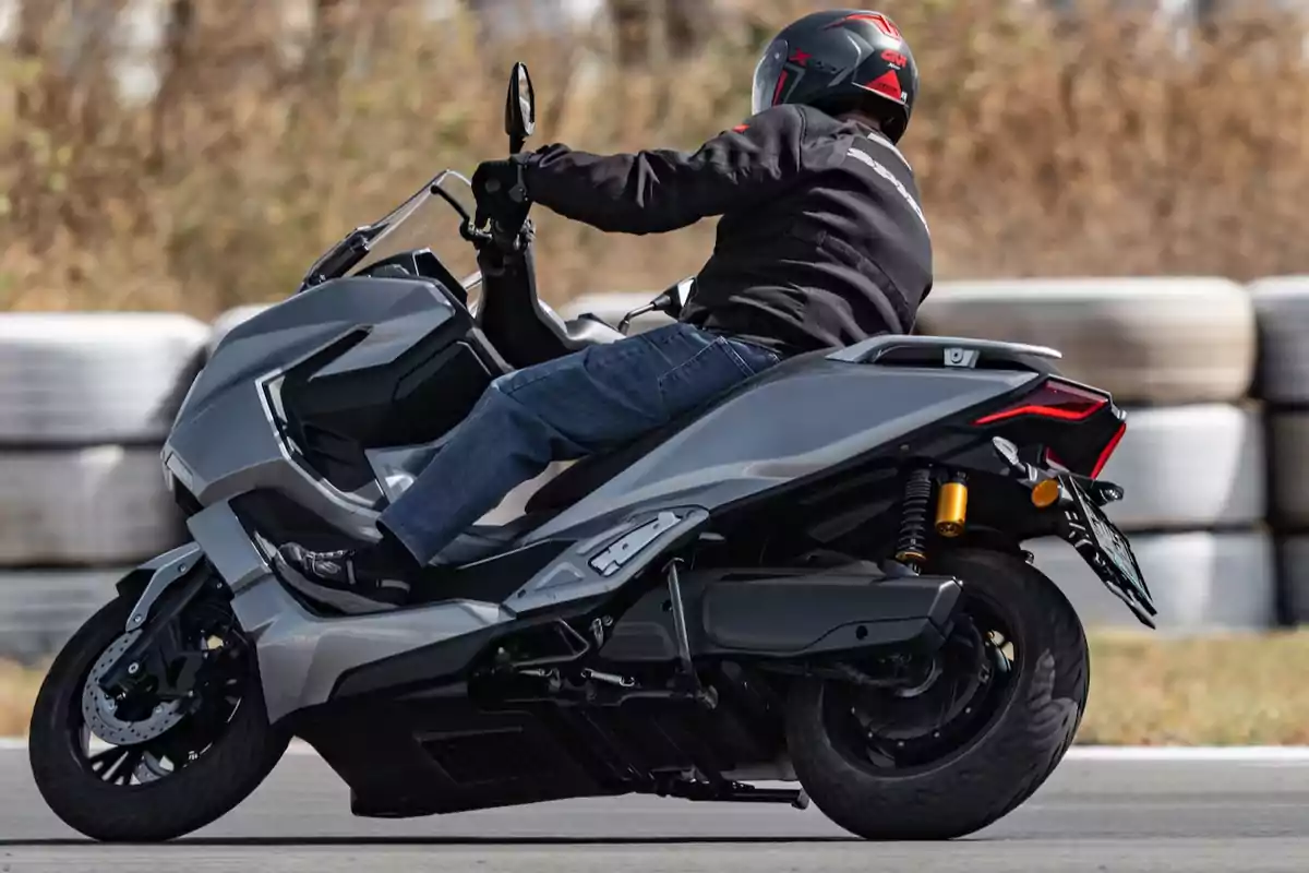 Persona conduciendo una motocicleta tipo scooter gris en una pista con fondo de llantas apiladas y vegetación seca. Persona conduciendo una motocicleta tipo scooter gris en una pista con fondo de llantas apiladas y vegetación seca.