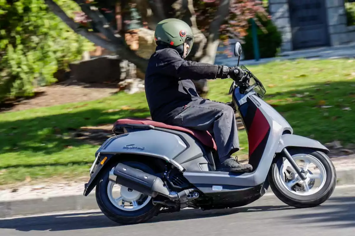 Person riding a gray scooter on a street with green areas in the background Person riding a gray scooter on a street with green areas in the background