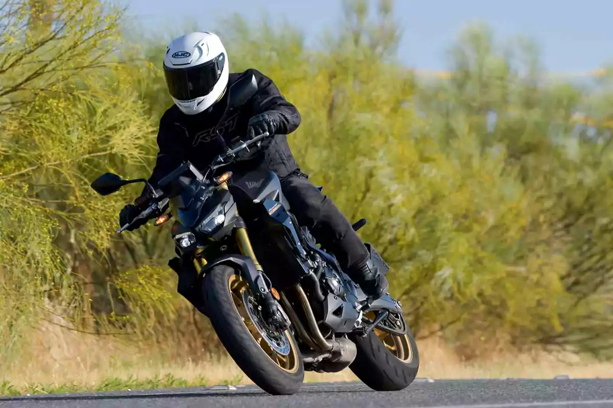 Persona con casco blanco y ropa negra conduciendo una motocicleta deportiva en una carretera rodeada de vegetación Persona con casco blanco y ropa negra conduciendo una motocicleta deportiva en una carretera rodeada de vegetación