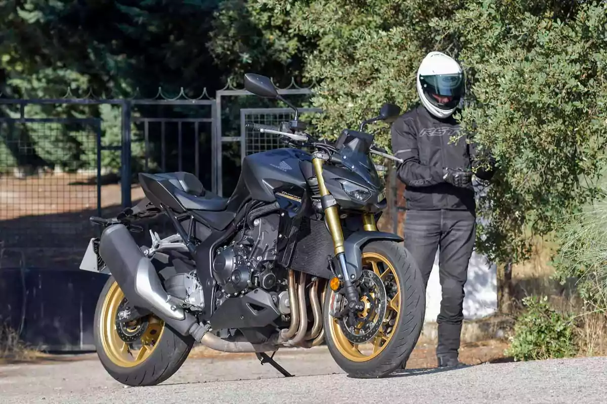 Motocicleta deportiva negra con detalles dorados estacionada en un camino junto a una persona con casco y ropa de protección para motociclista Motocicleta deportiva negra con detalles dorados estacionada en un camino junto a una persona con casco y ropa de protección para motociclista