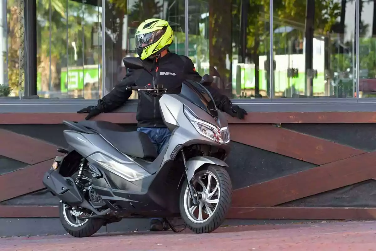 Persona con casco amarillo y chaqueta negra recargada en una moto tipo scooter gris frente a una ventana grande Persona con casco amarillo y chaqueta negra recargada en una moto tipo scooter gris frente a una ventana grande