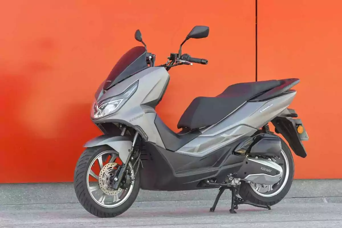 Motocicleta tipo scooter gris estacionada frente a una pared naranja Motocicleta tipo scooter gris estacionada frente a una pared naranja