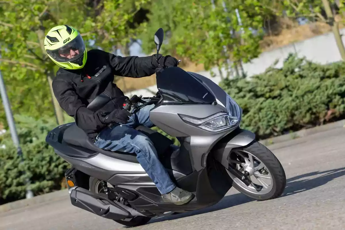 Persona conduciendo una motocicleta tipo scooter gris en una calle, usando casco amarillo y ropa de protección
