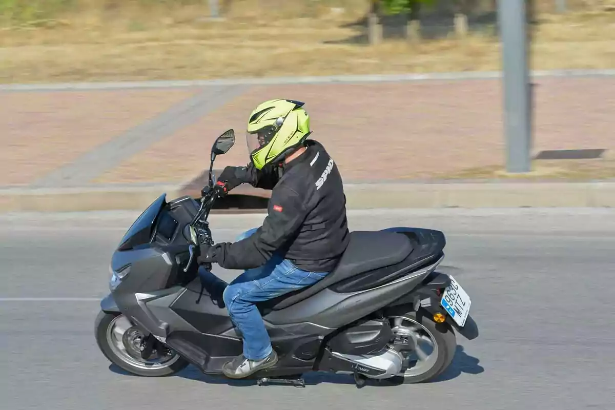 Persona conduciendo una motocicleta tipo scooter negra usando casco amarillo y chaqueta negra en una calle asfaltada Persona conduciendo una motocicleta tipo scooter negra usando casco amarillo y chaqueta negra en una calle asfaltada