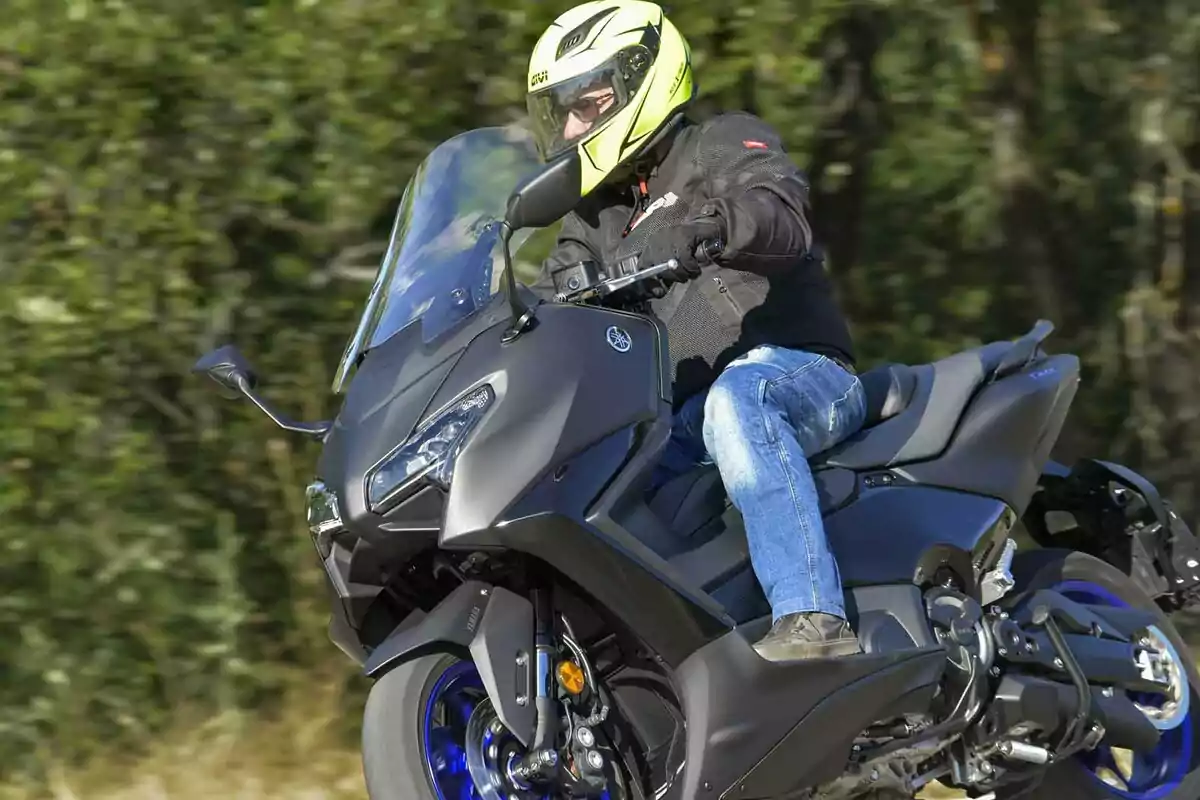 Persona conduciendo una motocicleta negra con casco amarillo y chaqueta oscura en un entorno al aire libre con vegetación de fondo Persona conduciendo una motocicleta negra con casco amarillo y chaqueta oscura en un entorno al aire libre con vegetación de fondo