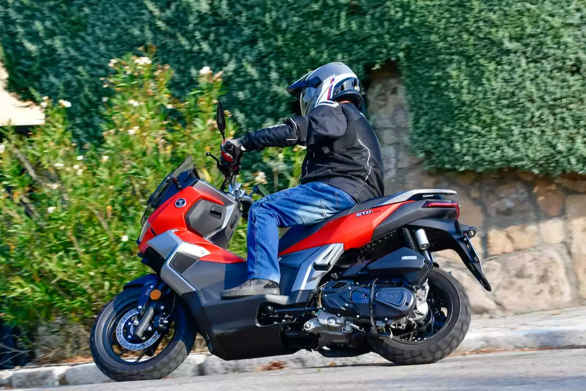 Person riding a red and black scooter down a street with vegetation and a stone wall in the background Person riding a red and black scooter down a street with vegetation and a stone wall in the background