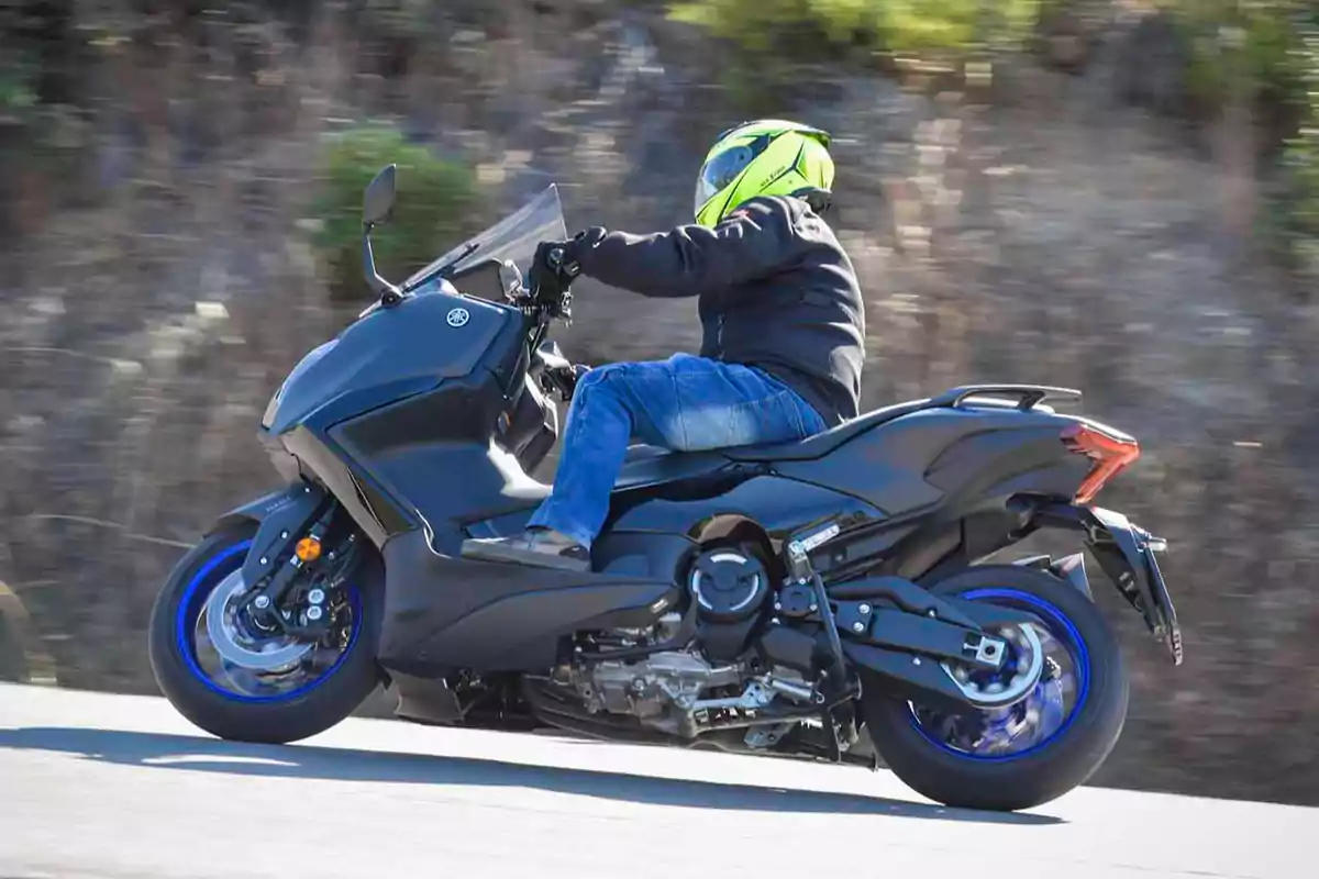 Persona conduciendo una motocicleta negra con detalles azules en una carretera, usando casco amarillo y chaqueta oscura Persona conduciendo una motocicleta negra con detalles azules en una carretera, usando casco amarillo y chaqueta oscura