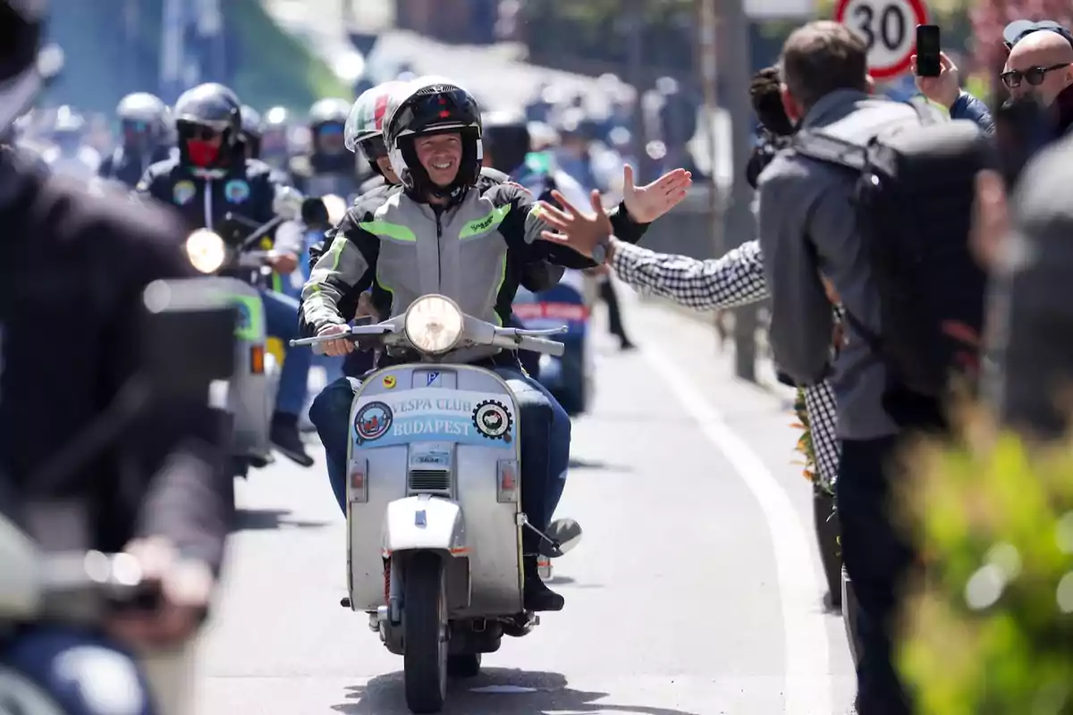 Un hombre sonriente conduce una Vespa en un desfile de motocicletas mientras choca la mano con una persona del público Un hombre sonriente conduce una Vespa en un desfile de motocicletas mientras choca la mano con una persona del público