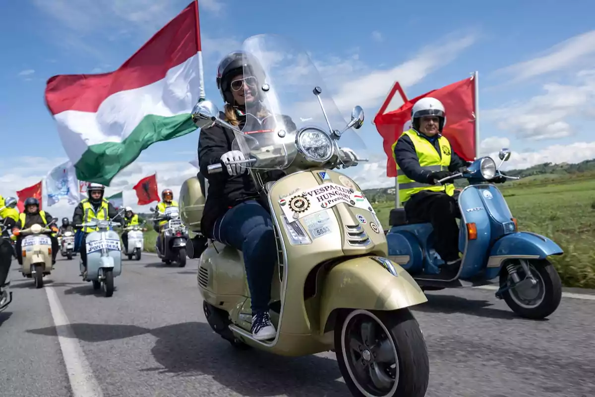Grupo de personas conduciendo motos Vespa en una carretera, algunas llevan banderas grandes y visten chalecos reflectantes, el cielo está despejado y el ambiente es festivo Grupo de personas conduciendo motos Vespa en una carretera, algunas llevan banderas grandes y visten chalecos reflectantes, el cielo está despejado y el ambiente es festivo