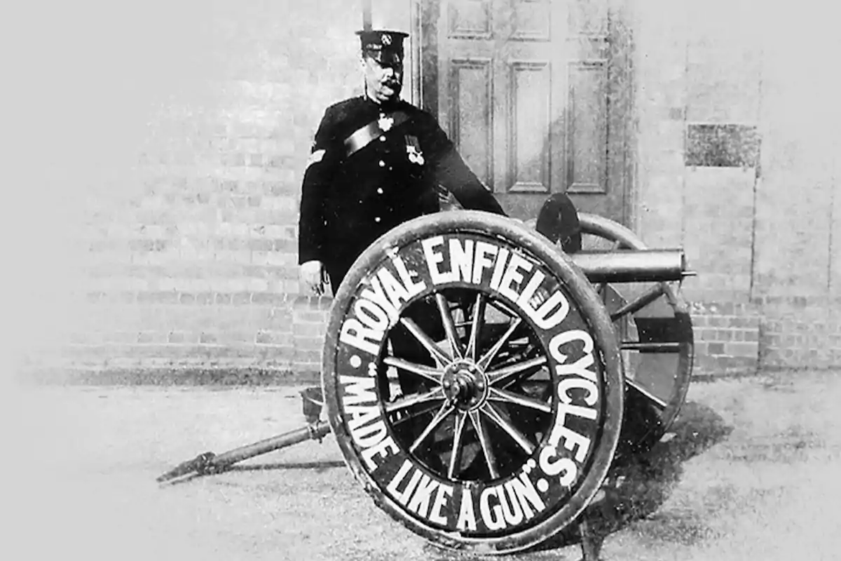 Fotografía antigua en blanco y negro de un hombre uniformado junto a un cañón cuya rueda tiene escrito Royal Enfield Cycles Made Like a Gun Fotografía antigua en blanco y negro de un hombre uniformado junto a un cañón cuya rueda tiene escrito Royal Enfield Cycles Made Like a Gun