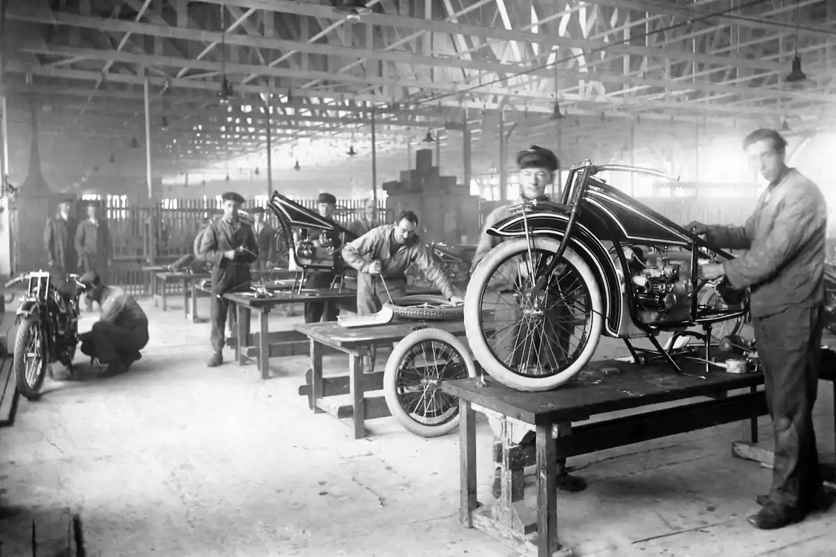 Varios hombres trabajan ensamblando motocicletas en una fábrica antigua con estructura de techo de vigas expuestas Varios hombres trabajan ensamblando motocicletas en una fábrica antigua con estructura de techo de vigas expuestas