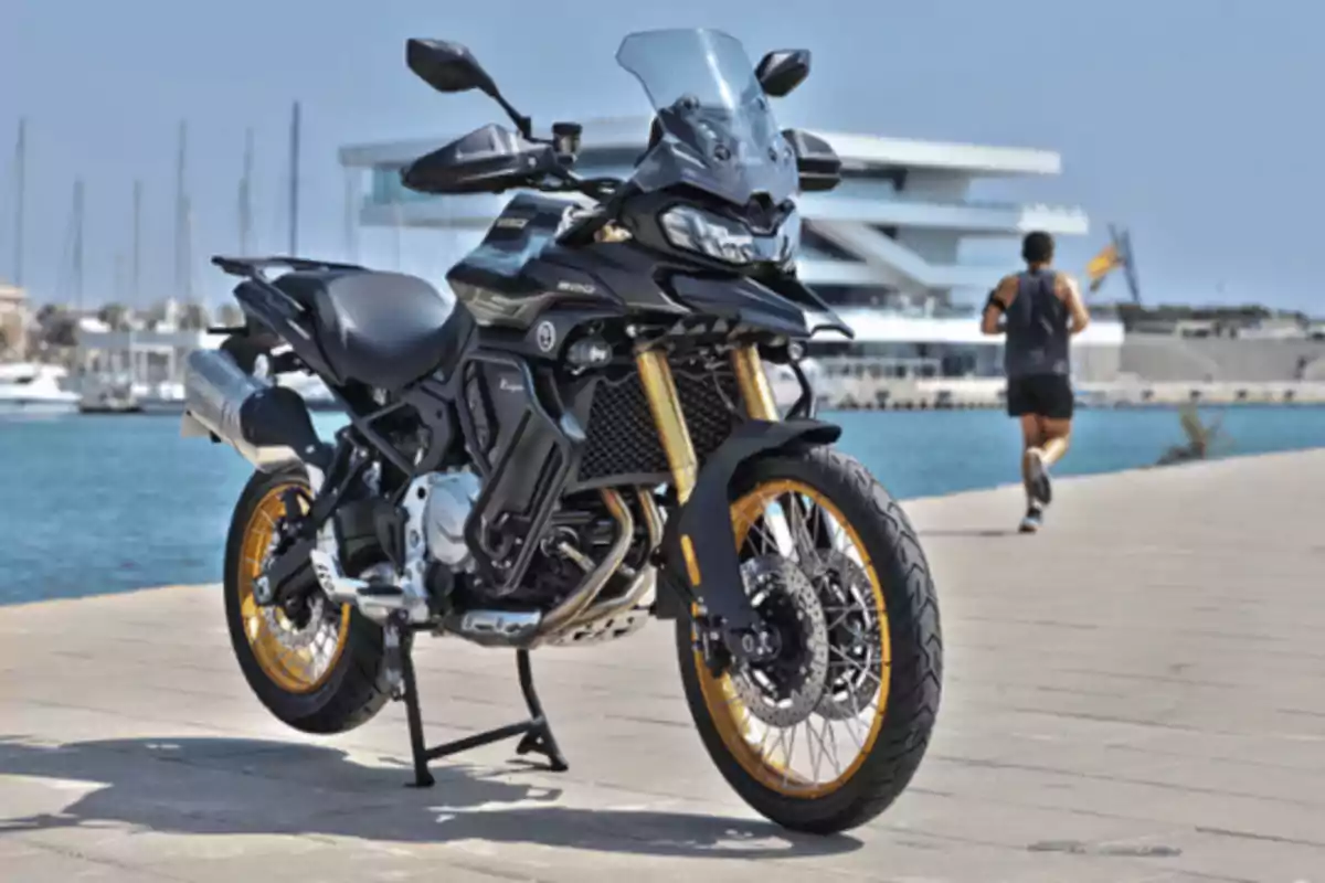 Motocicleta negra estacionada junto al mar en un muelle con una persona corriendo al fondo y barcos atracados Motocicleta negra estacionada junto al mar en un muelle con una persona corriendo al fondo y barcos atracados