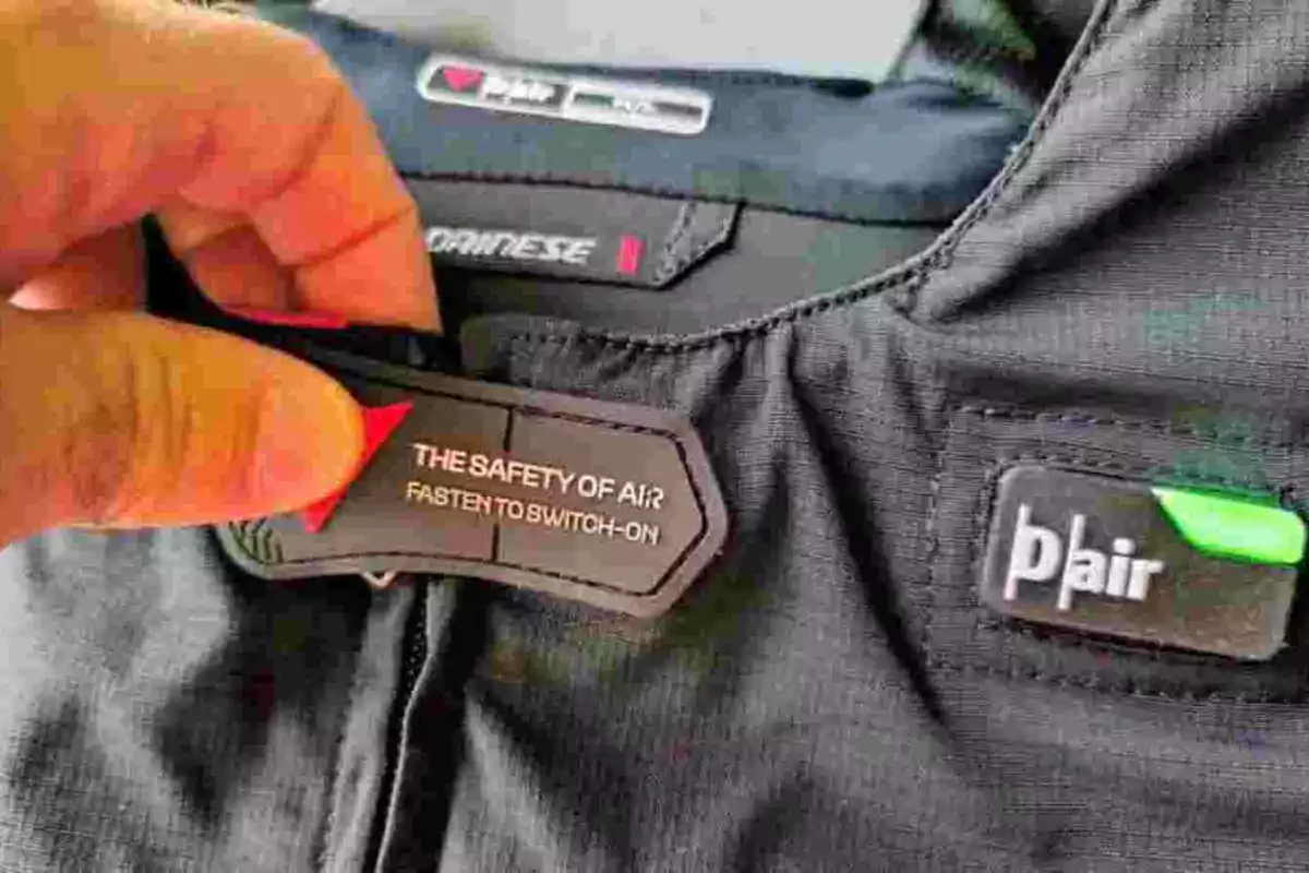 Una mano sostiene una etiqueta de un chaleco de motociclista con la inscripción THE SAFETY OF AIR FASTEN TO SWITCH-ON y el logo de Dainese y D-air visibles en la prenda. Una mano sostiene una etiqueta de un chaleco de motociclista con la inscripción THE SAFETY OF AIR FASTEN TO SWITCH-ON y el logo de Dainese y D-air visibles en la prenda.