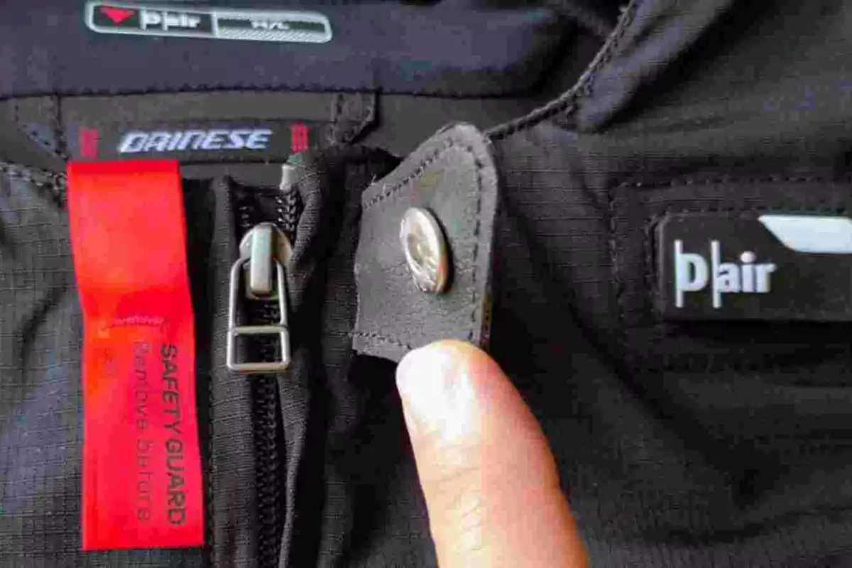 Una mano sostiene la solapa de un pantalón de motociclista negro con cierre, botón metálico y etiquetas visibles de las marcas Dainese y D-air Una mano sostiene la solapa de un pantalón de motociclista negro con cierre, botón metálico y etiquetas visibles de las marcas Dainese y D-air
