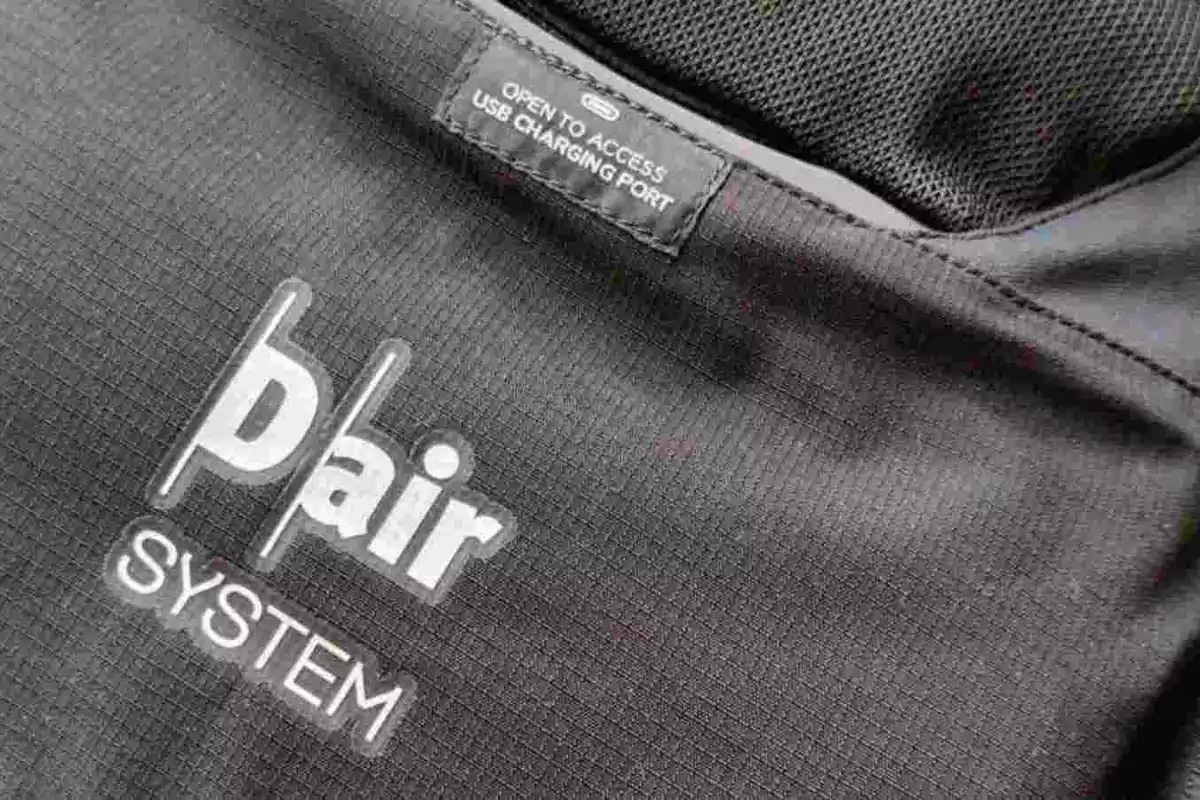 Detalle de una mochila negra con la inscripción Pair System y una etiqueta que indica acceso a puerto de carga USB Detalle de una mochila negra con la inscripción Pair System y una etiqueta que indica acceso a puerto de carga USB