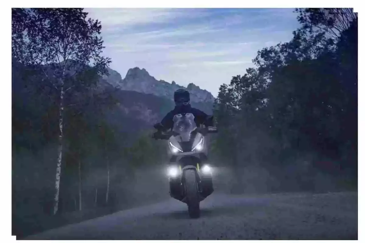 Motorcyclist riding a motorcycle with the lights on along a road surrounded by trees and mountains at sunset