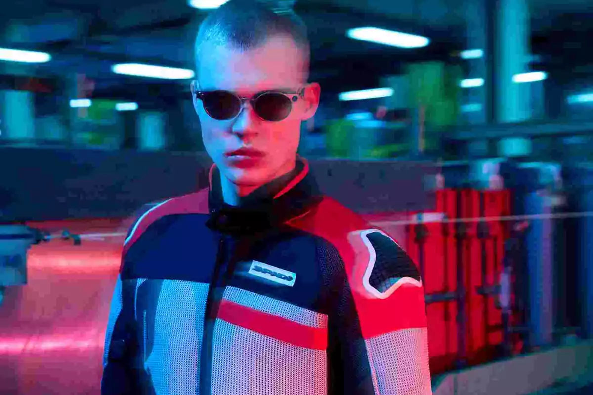 Joven con gafas de sol y chaqueta deportiva en un ambiente industrial iluminado con luces azules y rojas Joven con gafas de sol y chaqueta deportiva en un ambiente industrial iluminado con luces azules y rojas