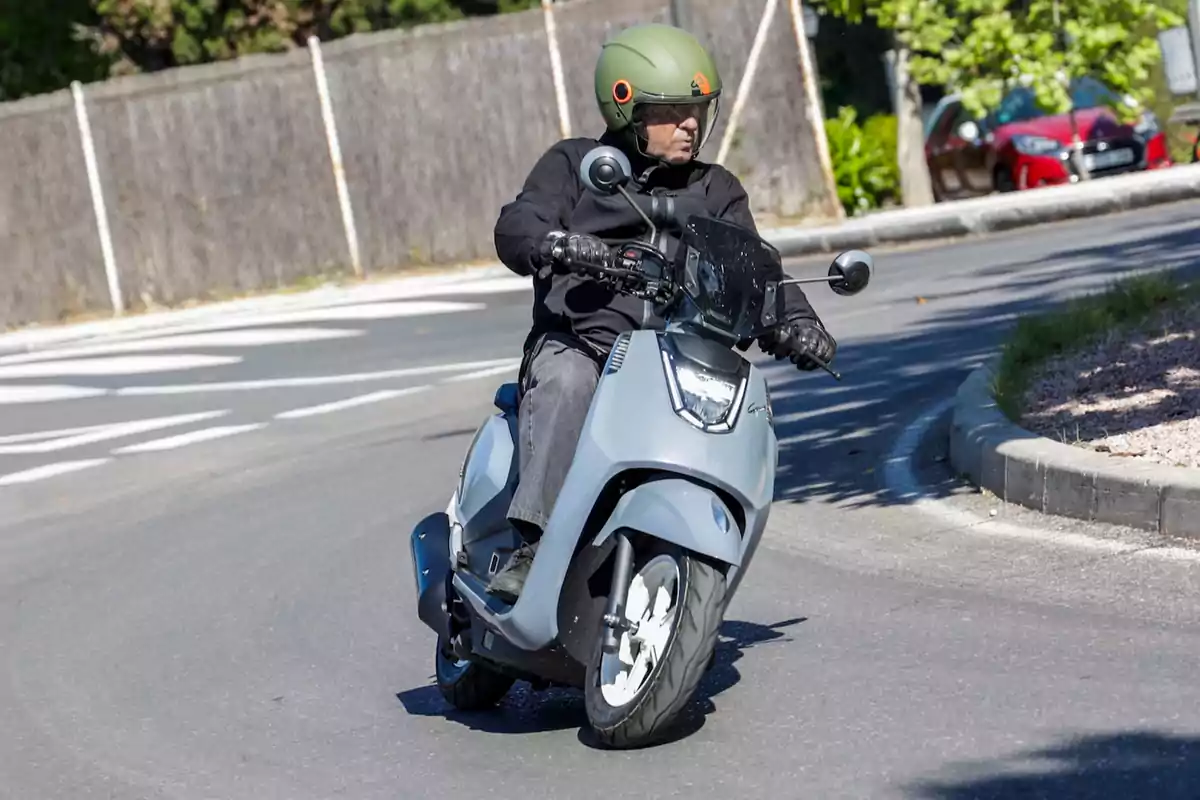 Persona conduciendo una moto tipo scooter gris en una curva de una calle asfaltada Persona conduciendo una moto tipo scooter gris en una curva de una calle asfaltada