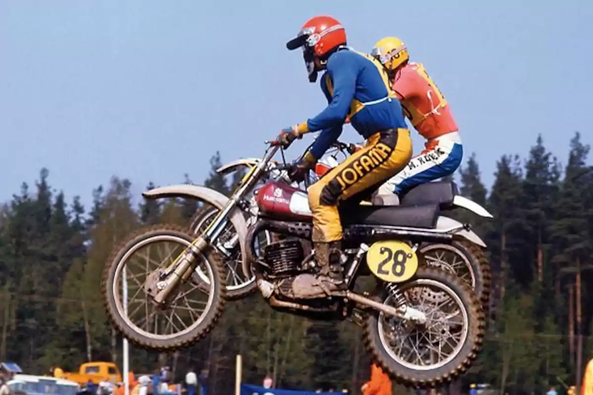 Dos motociclistas compiten en una carrera de motocross saltando por el aire en un entorno al aire libre rodeado de árboles Dos motociclistas compiten en una carrera de motocross saltando por el aire en un entorno al aire libre rodeado de árboles