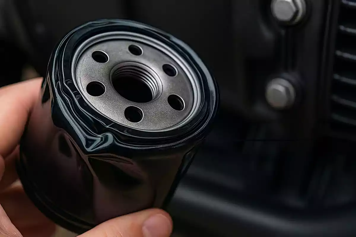 Persona sosteniendo un filtro de aceite de motor abollado frente a una parte del motor de un vehículo Persona sosteniendo un filtro de aceite de motor abollado frente a una parte del motor de un vehículo