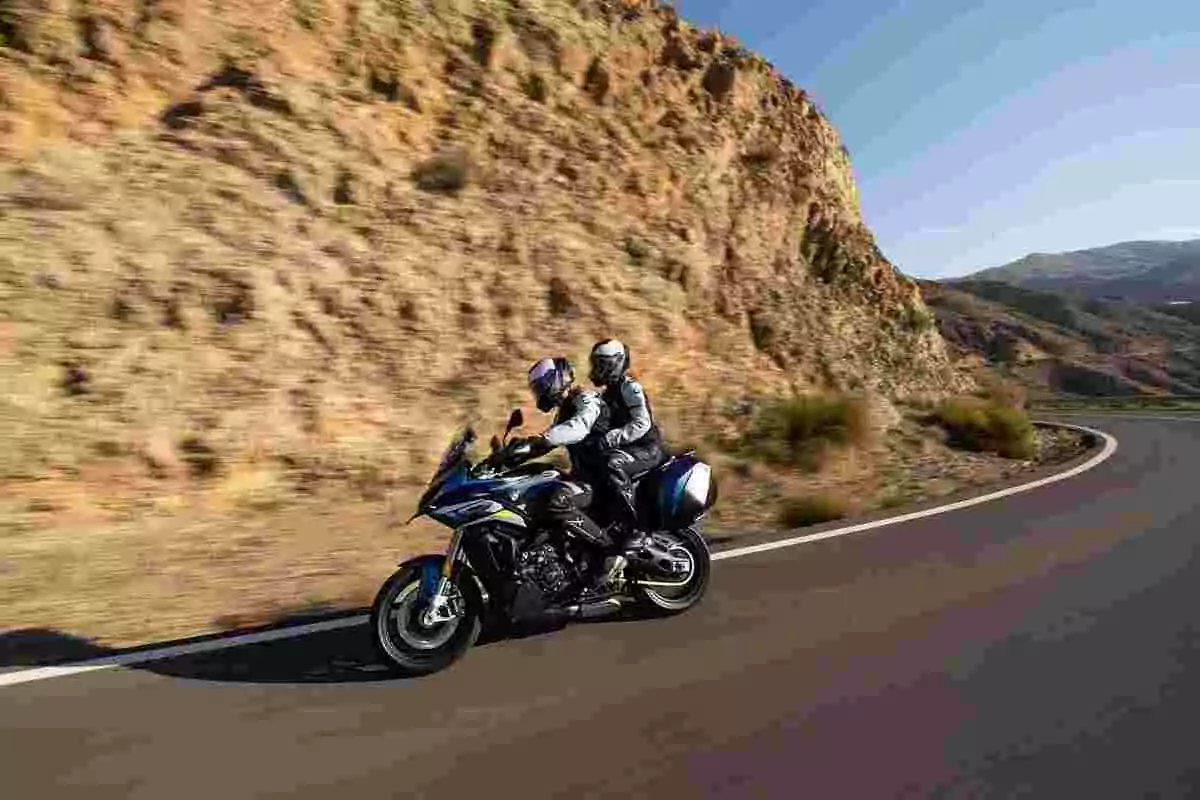 Dos personas con casco y equipo de protección viajan en motocicleta por una carretera curva junto a una montaña bajo un cielo despejado Dos personas con casco y equipo de protección viajan en motocicleta por una carretera curva junto a una montaña bajo un cielo despejado