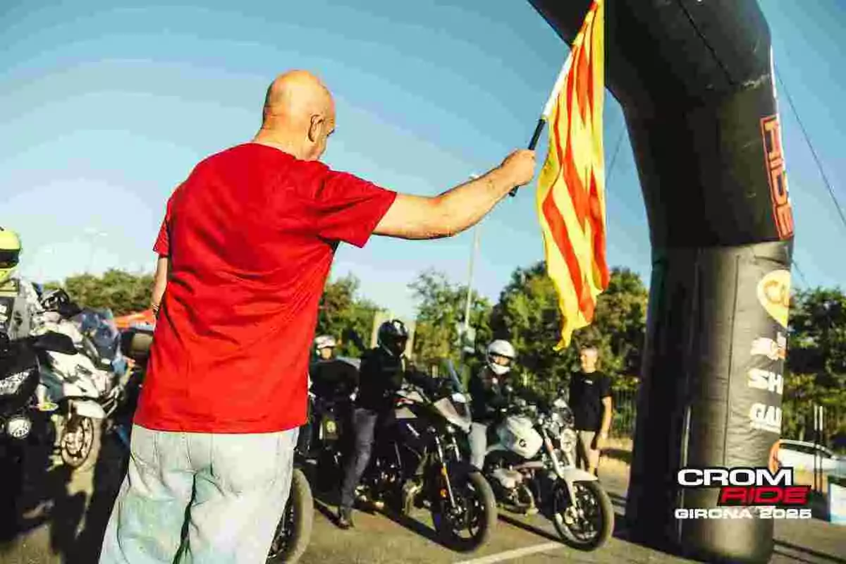 Un hombre con camiseta roja da la salida a un grupo de motoristas en un evento al aire libre ondeando una bandera amarilla con franjas rojas junto a un arco inflable que indica el inicio de la carrera Un hombre con camiseta roja da la salida a un grupo de motoristas en un evento al aire libre ondeando una bandera amarilla con franjas rojas junto a un arco inflable que indica el inicio de la carrera