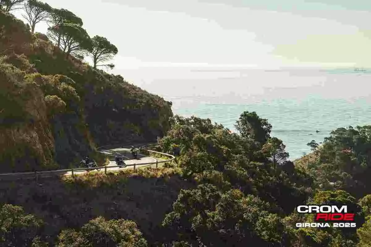 Motociclistas recorren una carretera costera rodeada de vegetación con vistas al mar y el logo de Crom Ride Girona 2025 en la esquina inferior derecha Motociclistas recorren una carretera costera rodeada de vegetación con vistas al mar y el logo de Crom Ride Girona 2025 en la esquina inferior derecha