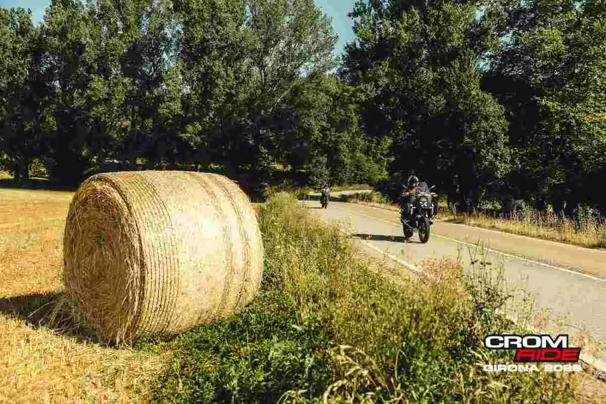 Un motociclista circula por una carretera rural junto a un campo con un gran rollo de paja y árboles al fondo, con el logo de Crom Ride Girona 2023 en la esquina inferior derecha. Un motociclista circula por una carretera rural junto a un campo con un gran rollo de paja y árboles al fondo, con el logo de Crom Ride Girona 2023 en la esquina inferior derecha.