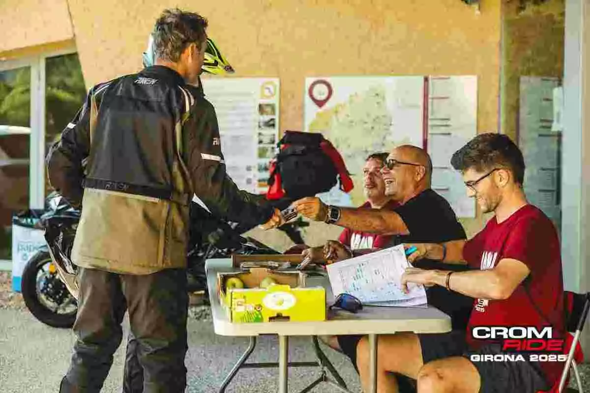 Un motociclista entrega un documento a un grupo de tres personas sentadas en una mesa de registro durante un evento llamado Crom Ride Girona 2026 Un motociclista entrega un documento a un grupo de tres personas sentadas en una mesa de registro durante un evento llamado Crom Ride Girona 2026