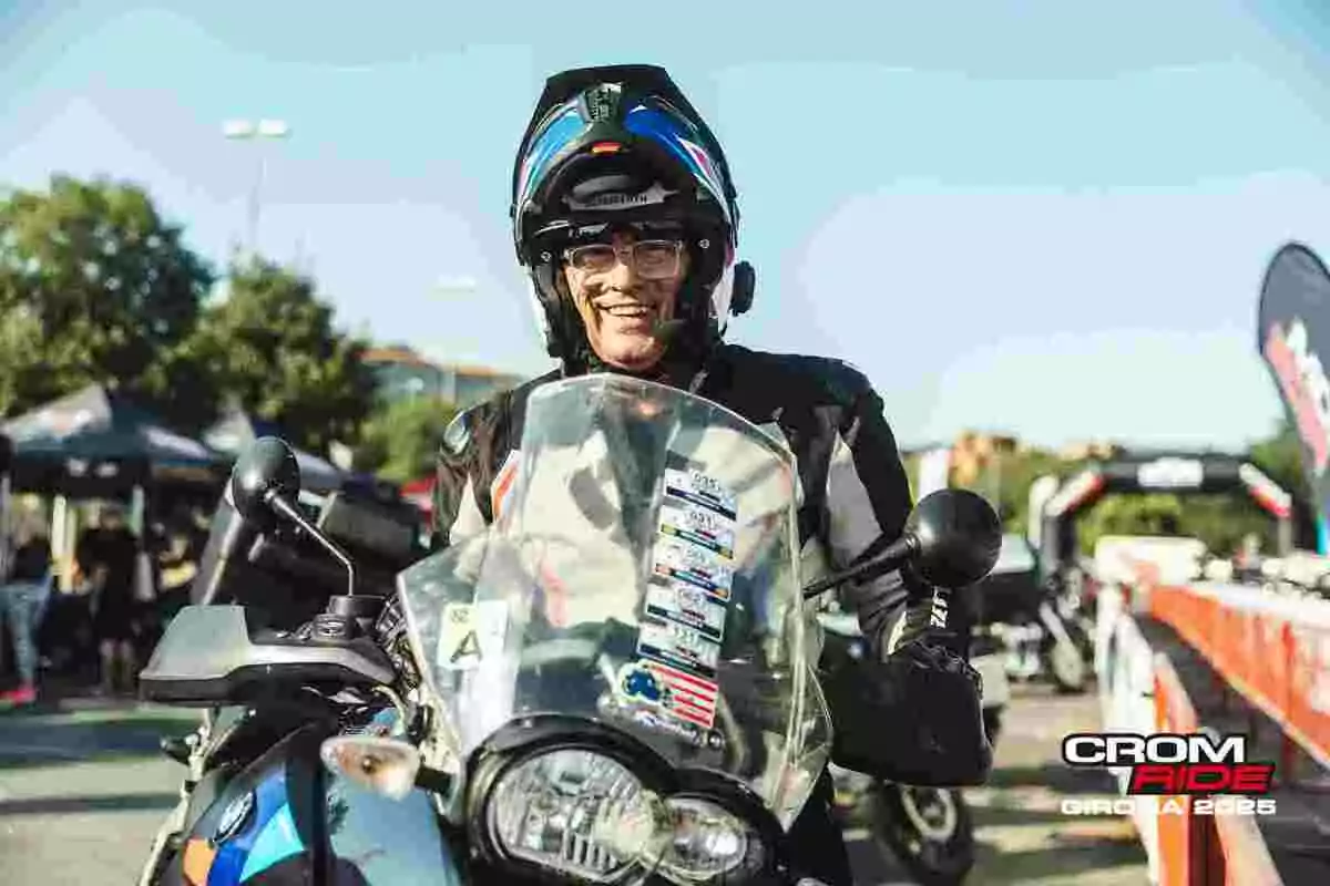 Persona sonriente con casco y gafas montada en una motocicleta durante un evento al aire libre con ambiente soleado y carpas de fondo Persona sonriente con casco y gafas montada en una motocicleta durante un evento al aire libre con ambiente soleado y carpas de fondo