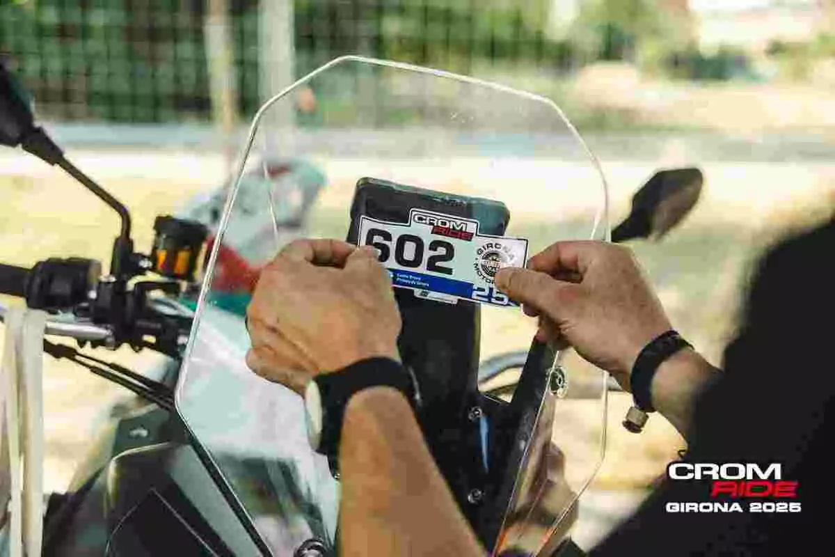 Manos colocando una pegatina de evento en la pantalla de una motocicleta con el logo de CROM RIDE GIRONA 2025 visible en la esquina Manos colocando una pegatina de evento en la pantalla de una motocicleta con el logo de CROM RIDE GIRONA 2025 visible en la esquina