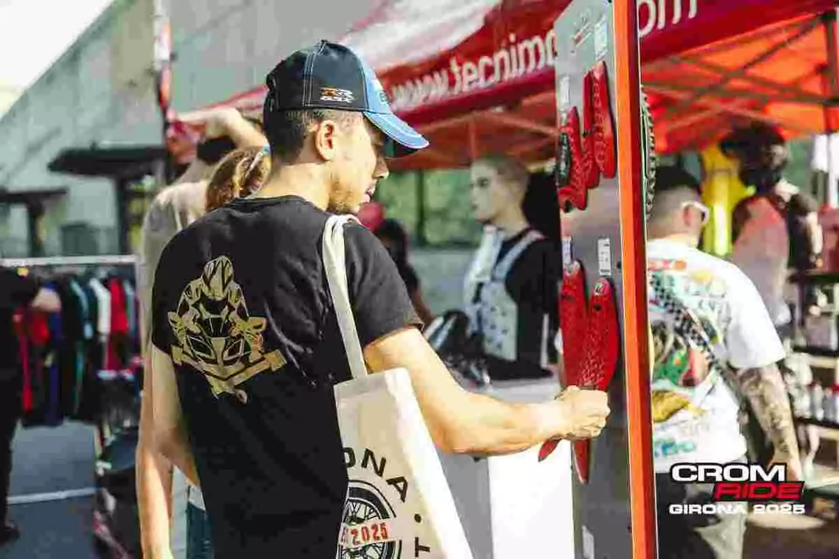 Hombre con gorra y camiseta negra observa productos en un stand al aire libre durante un evento de motociclismo en Girona 2025 Hombre con gorra y camiseta negra observa productos en un stand al aire libre durante un evento de motociclismo en Girona 2025