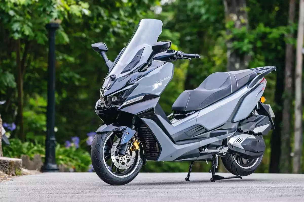 Motocicleta tipo scooter gris estacionada en una calle rodeada de árboles y vegetación Motocicleta tipo scooter gris estacionada en una calle rodeada de árboles y vegetación