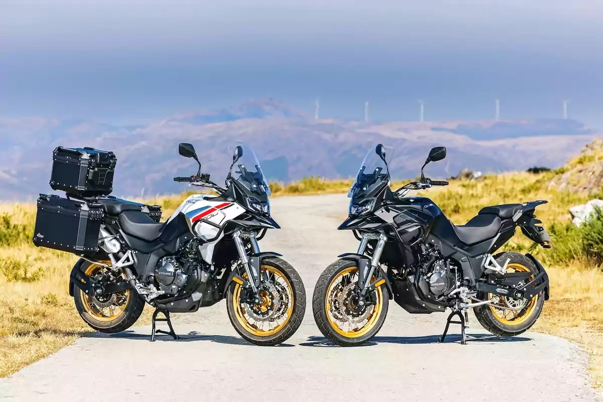 Dos motocicletas tipo touring están estacionadas una frente a la otra en un camino al aire libre, con un paisaje montañoso y molinos de viento al fondo. Dos motocicletas tipo touring están estacionadas una frente a la otra en un camino al aire libre, con un paisaje montañoso y molinos de viento al fondo.