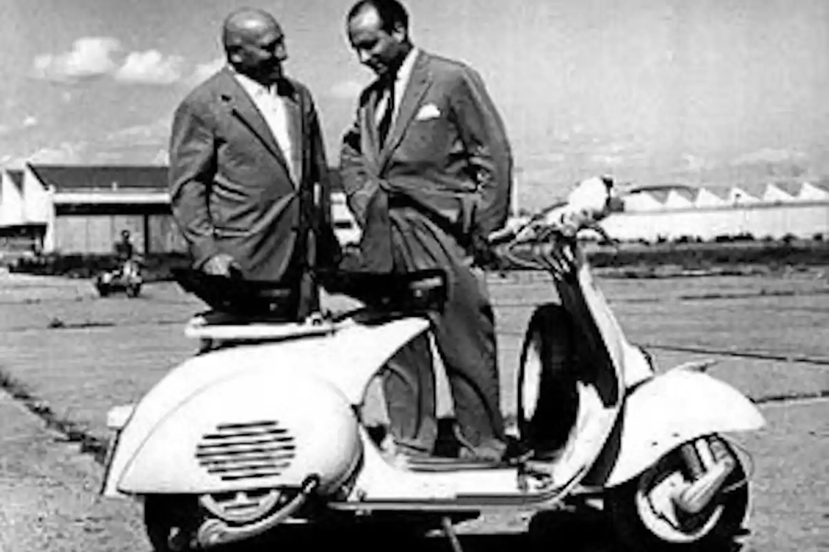 Dos hombres de traje conversan junto a una motoneta clásica en blanco y negro en un entorno industrial Dos hombres de traje conversan junto a una motoneta clásica en blanco y negro en un entorno industrial