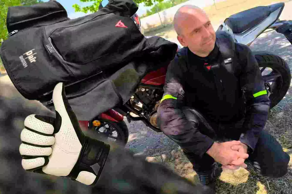 Hombre con chaqueta de motociclista junto a una moto, una chaqueta negra sobre la moto y un guante blanco haciendo señal de aprobación en primer plano