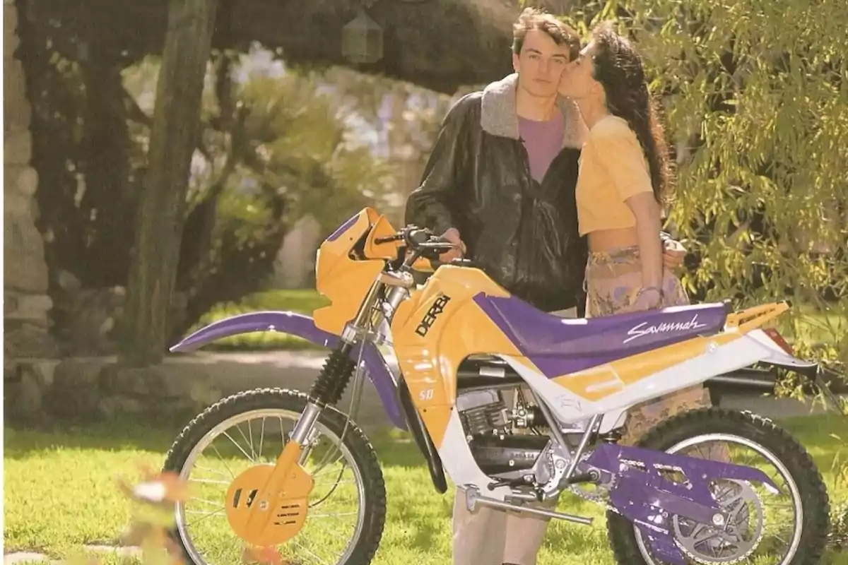A young couple next to a yellow and purple Derbi motorcycle in an outdoor setting with grass and trees
