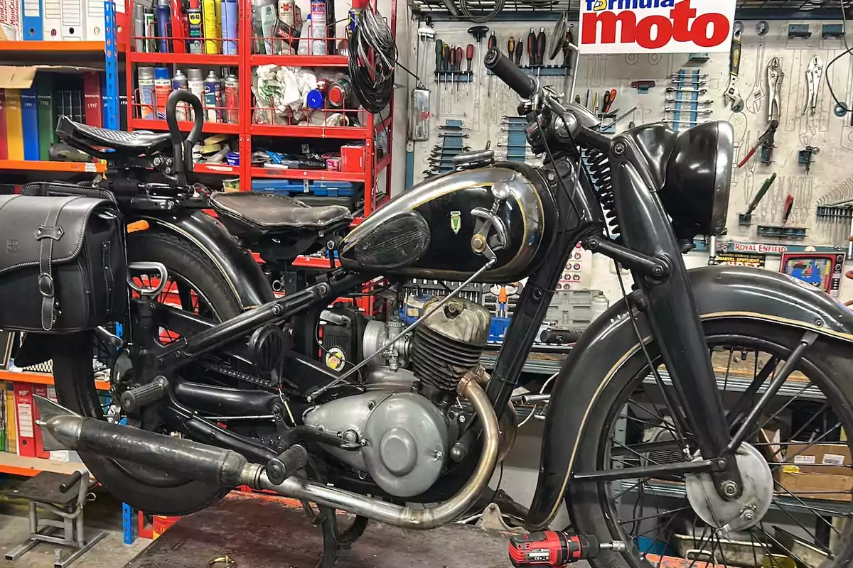 Motocicleta clásica negra en un taller mecánico con herramientas y estanterías al fondo Motocicleta clásica negra en un taller mecánico con herramientas y estanterías al fondo
