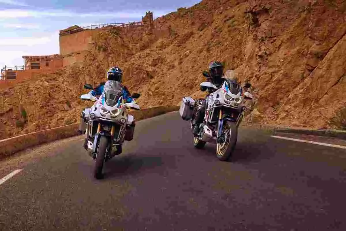 Dos motociclistas circulan por una carretera de montaña junto a un paisaje rocoso y edificaciones de fondo Dos motociclistas circulan por una carretera de montaña junto a un paisaje rocoso y edificaciones de fondo