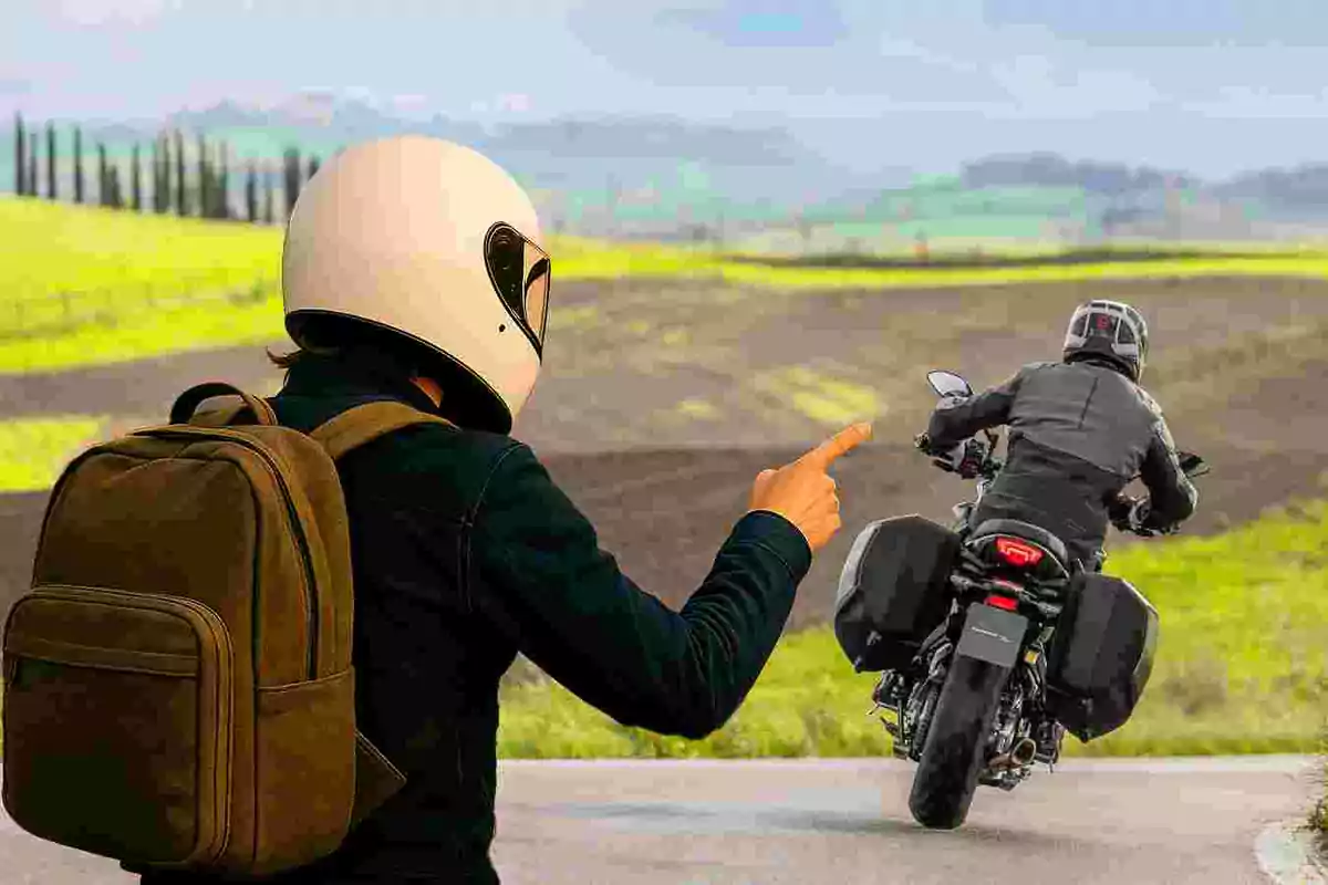 Persona con casco y mochila señalando a un motociclista que se aleja por un camino rural rodeado de campos verdes