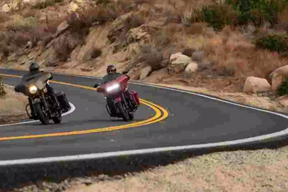 Dos motociclistas manejan por una carretera curva en una zona montañosa con vegetación seca y rocas.