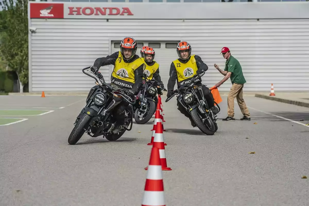 Tres persones amb armilles grogues i cascos taronges condueixen motocicletes Honda entre cons en un circuit d’entrenament mentre un instructor observa al fons.