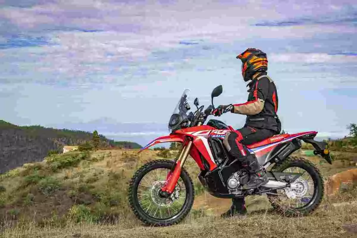 Persona con casco y equipo de protección montando una motocicleta Honda roja en un paisaje de campo abierto con colinas y cielo parcialmente nublado Persona con casco y equipo de protección montando una motocicleta Honda roja en un paisaje de campo abierto con colinas y cielo parcialmente nublado
