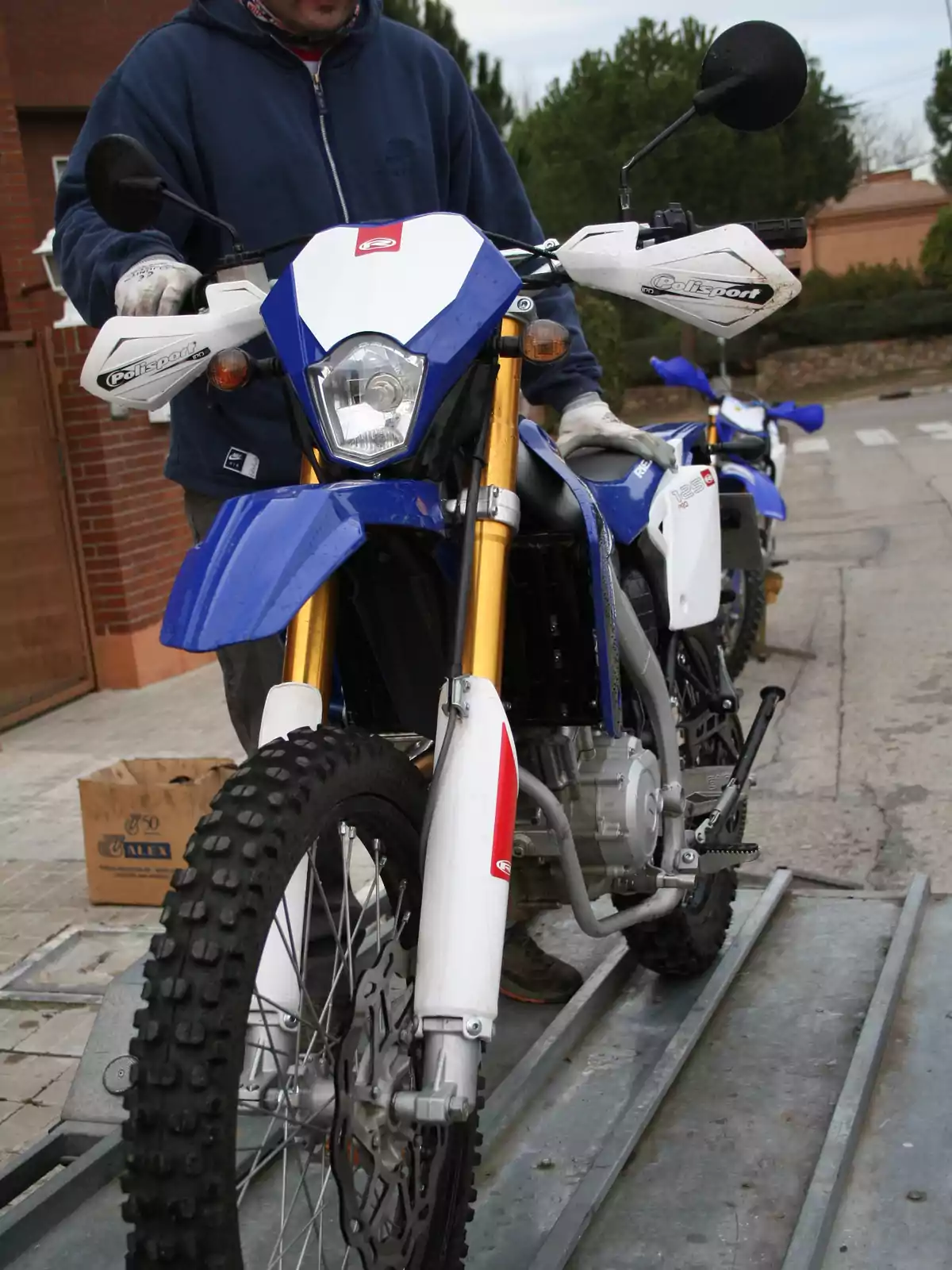 Persona con sudadera azul y guantes sosteniendo una motocicleta de enduro azul y blanca sobre una rampa en una calle residencial Persona con sudadera azul y guantes sosteniendo una motocicleta de enduro azul y blanca sobre una rampa en una calle residencial