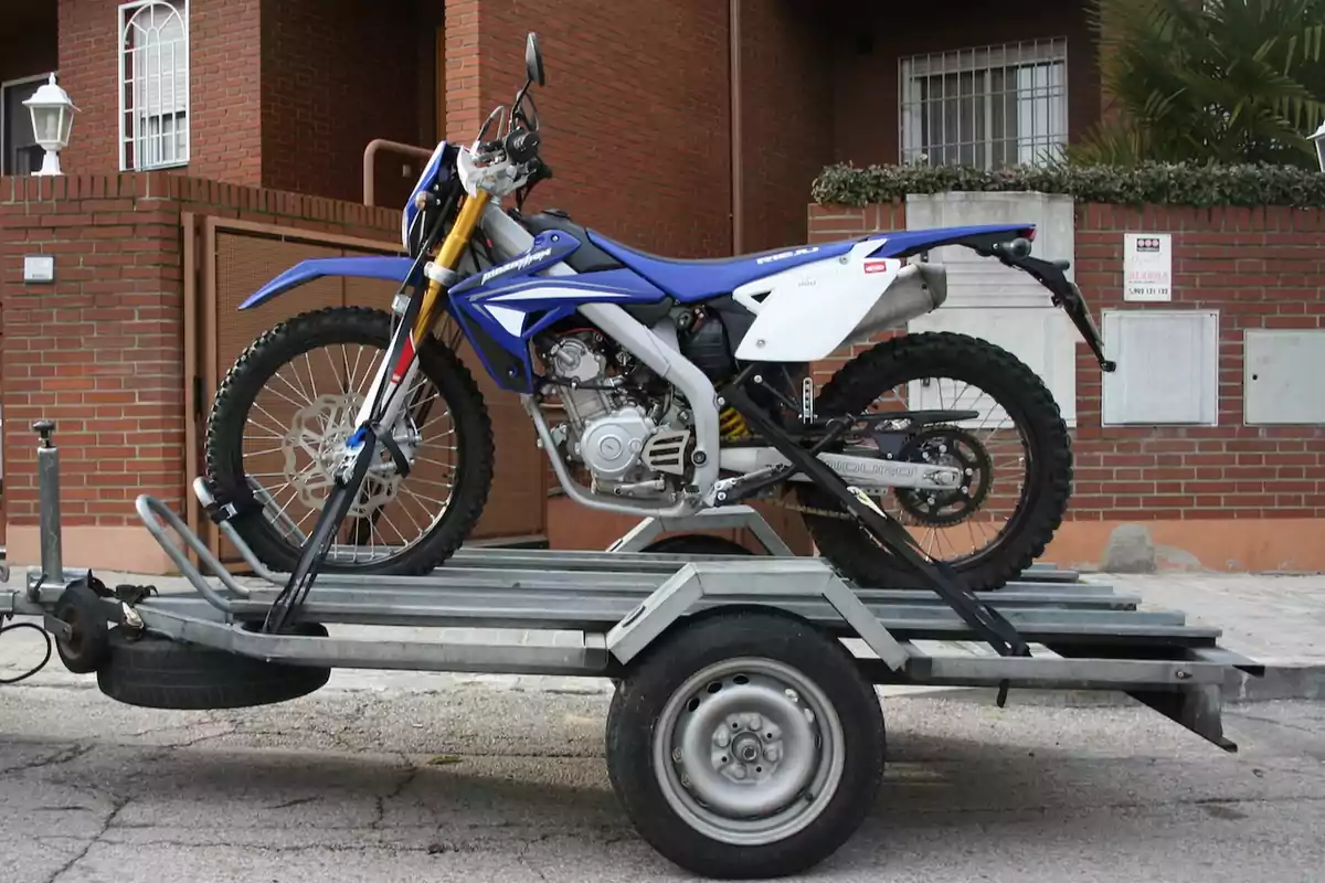 Motocicleta de motocross azul asegurada sobre un remolque metálico estacionado frente a una casa de ladrillo Motocicleta de motocross azul asegurada sobre un remolque metálico estacionado frente a una casa de ladrillo