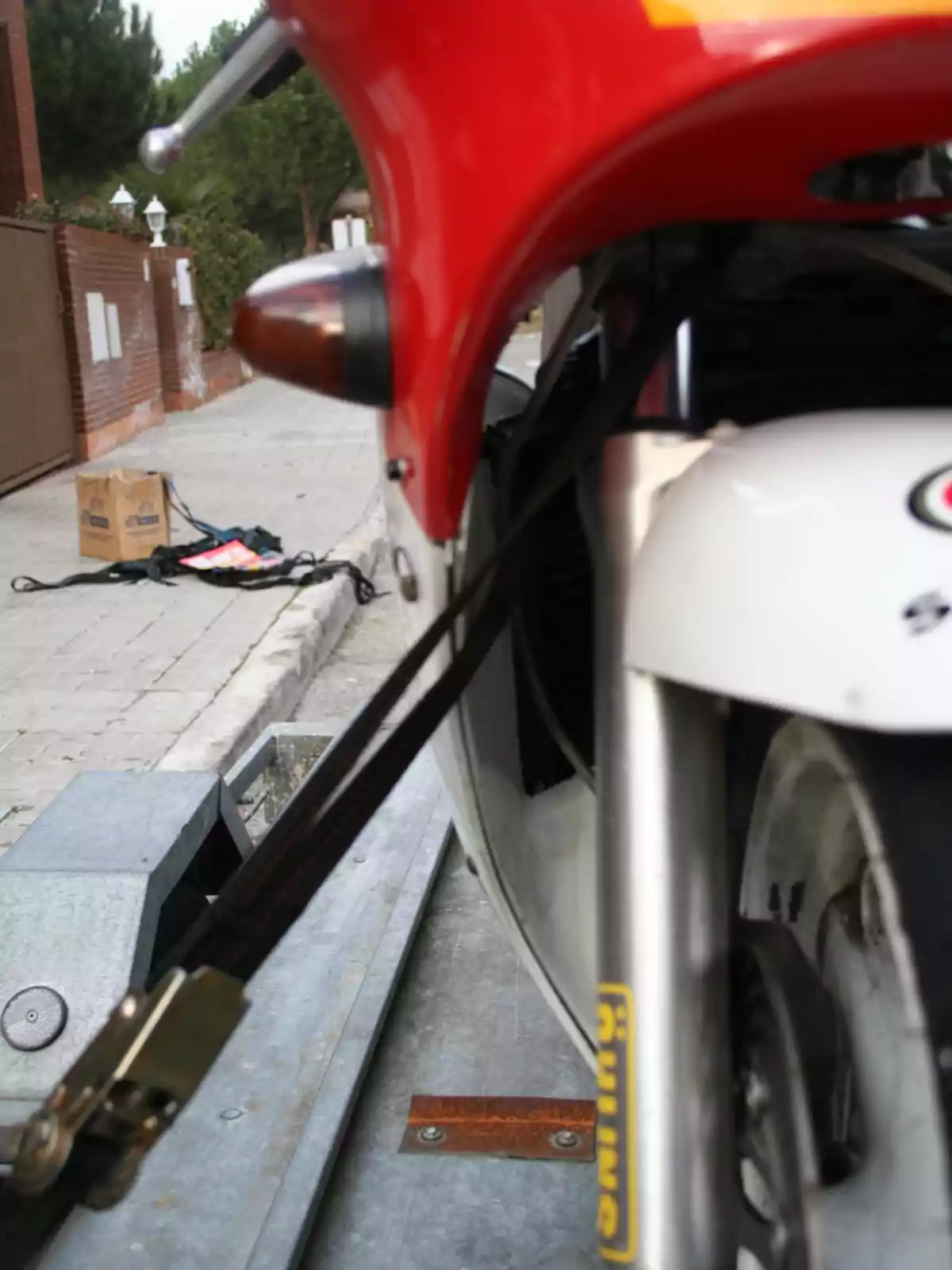 Motocicleta roja asegurada con una correa en una plataforma metálica, con una caja de cartón y objetos en la acera al fondo Motocicleta roja asegurada con una correa en una plataforma metálica, con una caja de cartón y objetos en la acera al fondo