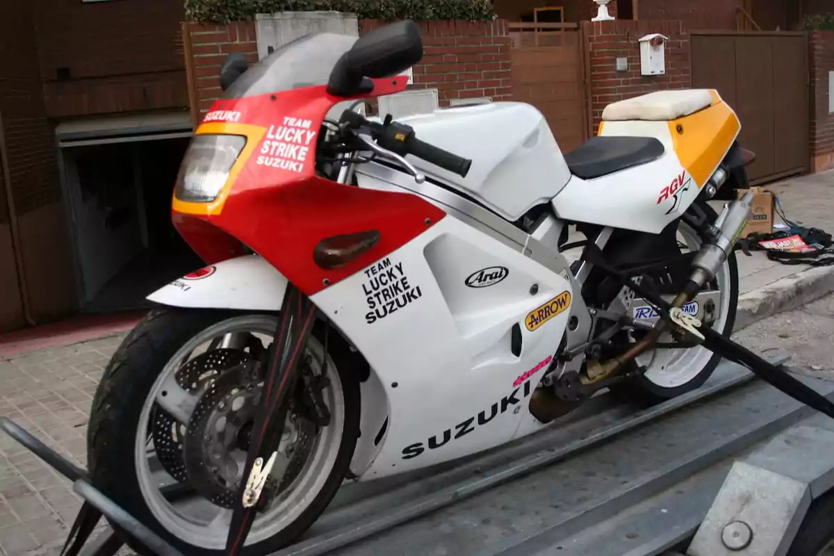 Motocicleta deportiva Suzuki RGV con carenado blanco, rojo y amarillo, decorada con logotipos de patrocinadores y asegurada sobre un remolque en una calle residencial