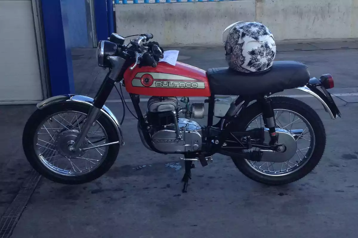 Motocicleta clásica Bultaco de color rojo estacionada junto a una pared con un casco blanco decorado sobre el asiento Motocicleta clásica Bultaco de color rojo estacionada junto a una pared con un casco blanco decorado sobre el asiento