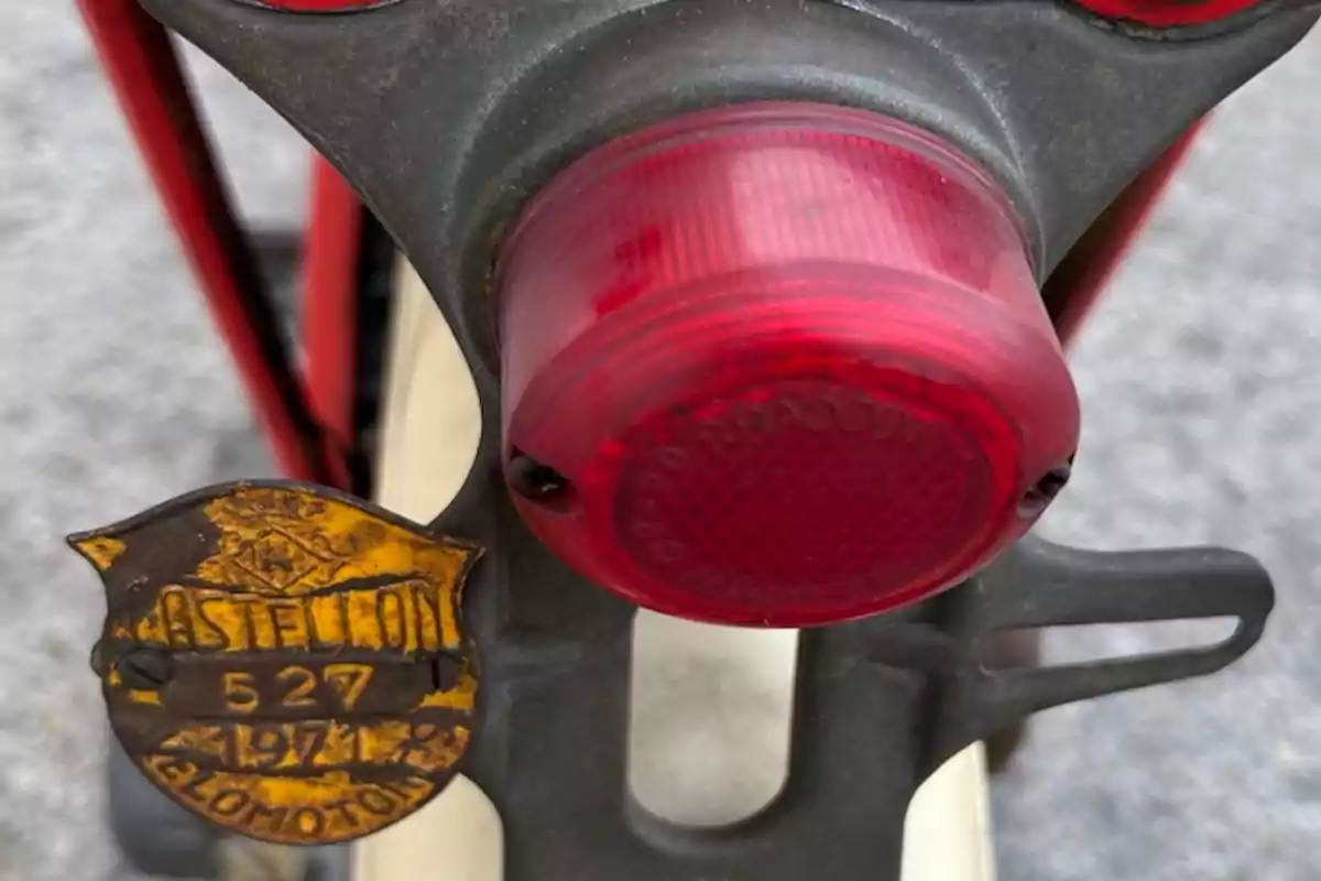 Detalle de la parte trasera de una bicicleta antigua con una luz roja y una placa metálica oxidada que dice Castellon 527 1971 Velomotor Detalle de la parte trasera de una bicicleta antigua con una luz roja y una placa metálica oxidada que dice Castellon 527 1971 Velomotor