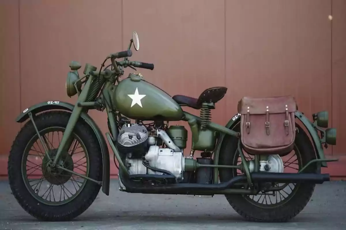 Motocicleta militar antigua de color verde oliva con una estrella blanca en el tanque y una alforja marrón en la parte trasera estacionada frente a una pared marrón Motocicleta militar antigua de color verde oliva con una estrella blanca en el tanque y una alforja marrón en la parte trasera estacionada frente a una pared marrón